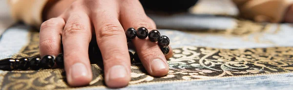 Cropped view of man holding prayer beads while praying on rug at home, banner — Foto stock