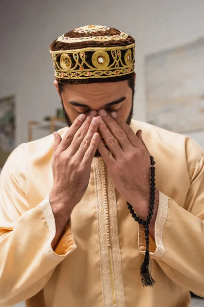 Young muslim man with prayer beads holding hands near face at home — Photo de stock