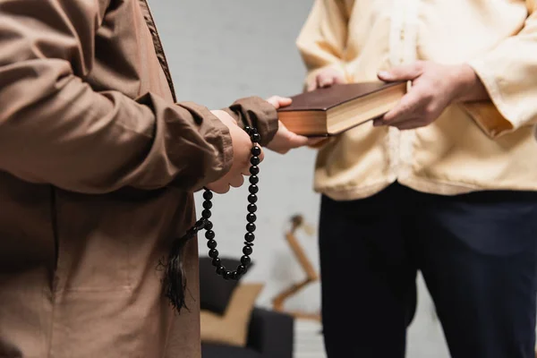 Cropped view of blurred muslim dad giving koran to son with prayer beads at home — Stock Photo