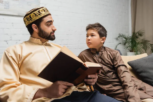 Padre musulmán sosteniendo el libro y mirando a su hijo en el sofá en casa - foto de stock