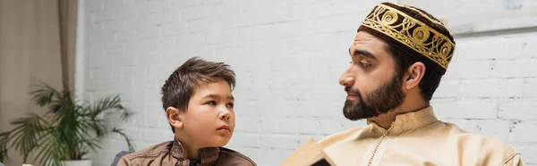 Middle eastern man looking at son near blurred book at home, banner — Stockfoto