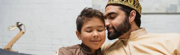 Padre musulmán sonriendo cerca de hijo preadolescente en casa, bandera - foto de stock