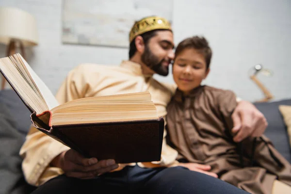 Book in hand of muslim man hugging son on couch at home — Foto stock