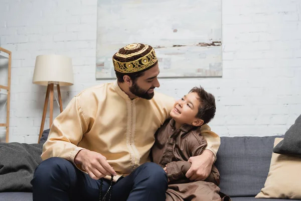 Homme musulman avec des perles de prière étreignant son fils sur le canapé à la maison — Photo de stock