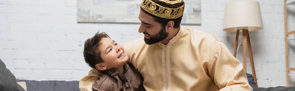 Sonriente musulmán hombre abrazando preadolescente hijo en casa, pancarta - foto de stock