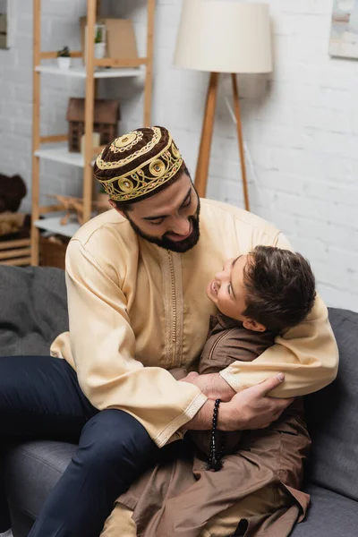 Cheerful muslim man with prayer beads hugging son on couch at home — Stock Photo