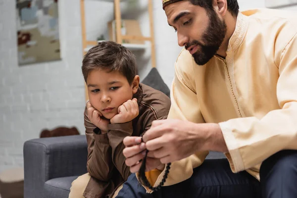 Homme musulman tenant des perles de prière et parlant à son fils sur le canapé à la maison — Photo de stock
