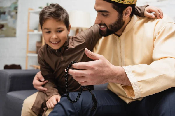 Hombre musulmán sosteniendo cuentas de oración y abrazando a su hijo borroso en casa - foto de stock