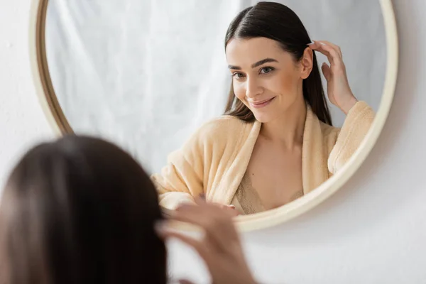Joven morena mujer ajustando el pelo y sonriendo mientras mira el espejo en el baño - foto de stock