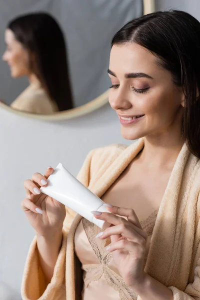 Heureuse jeune femme en peignoir regardant tube avec de la crème dans la salle de bain — Photo de stock