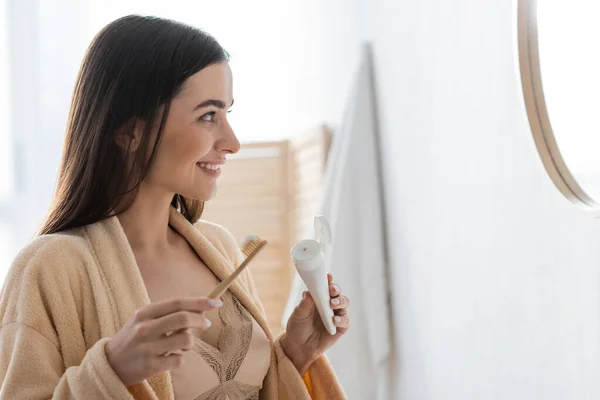 Side view of happy young woman holding tube with toothpaste and toothbrush while looking at mirror — Stock Photo