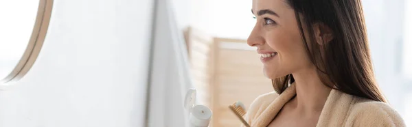 Side view of happy young woman holding tube with toothpaste and toothbrush while looking at mirror, banner — Stock Photo
