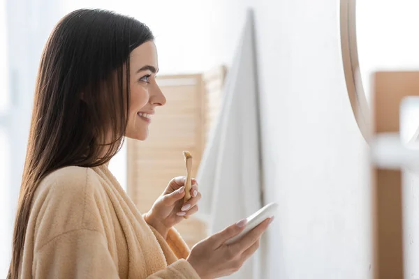 Side view of cheerful young woman holding toothpaste and toothbrush while looking at mirror — Stock Photo