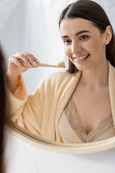 Young and cheerful woman holding toothbrush and looking at mirror in bathroom — Stockfoto