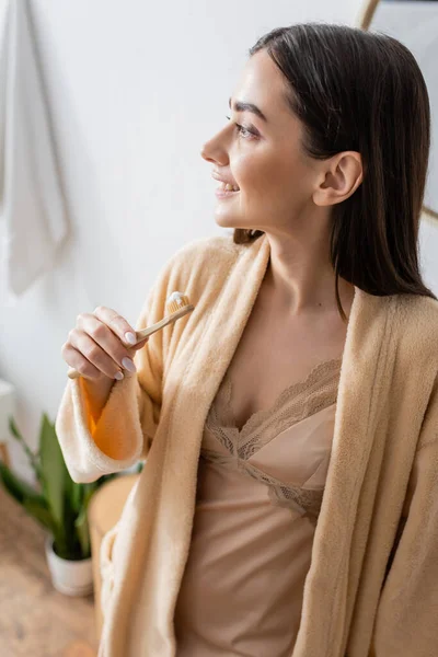 Young and cheerful woman in bathrobe holding toothbrush with toothpaste in bathroom — Stock Photo