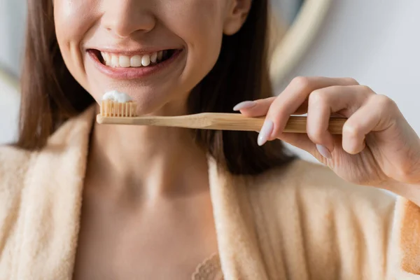Vue partielle de la femme souriante en peignoir tenant brosse à dents avec dentifrice dans la salle de bain — Photo de stock