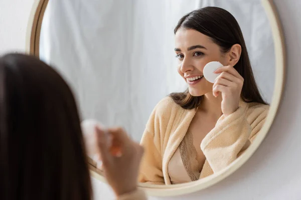 Happy young woman in bathrobe cleansing face with cotton pad in bathroom — Photo de stock