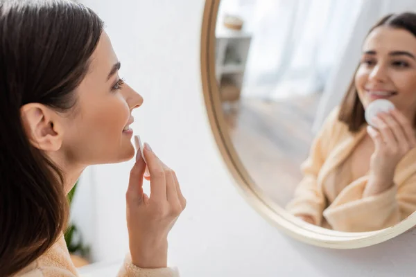 Smiling young woman in bathrobe cleansing face with cotton pad and looking at mirror in bathroom — Stock Photo