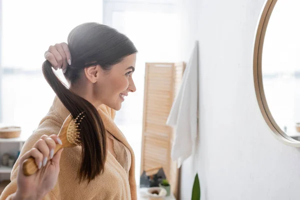 Side view of happy young woman brushing shiny hair and looking at mirror in bathroom — Stock Photo