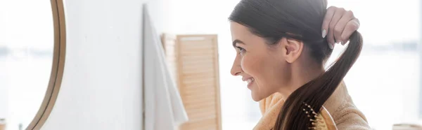 Side view of happy young woman brushing shiny hair and looking at mirror in bathroom, banner — Stock Photo