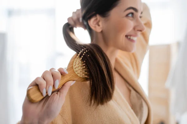 Smiling and blurred young woman brushing shiny hair in bathroom — Stock Photo