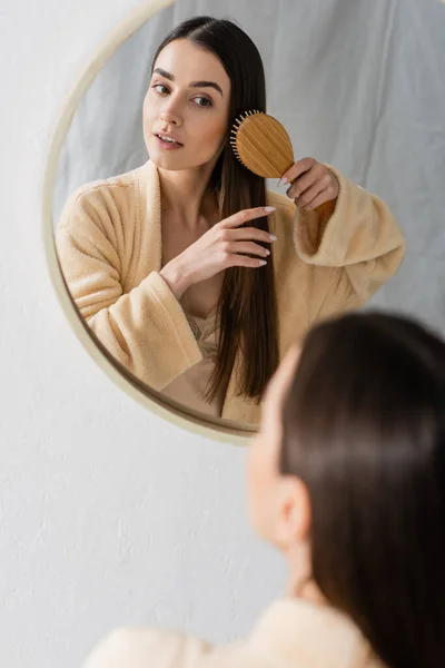 Reflection of brunette young woman brushing shiny hair and looking at mirror in bathroom — Stock Photo
