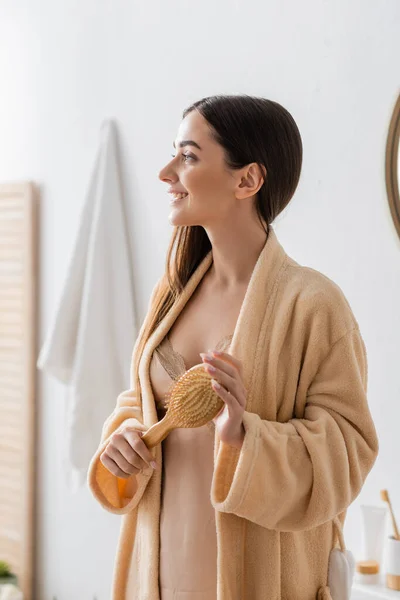 Side view of joyful young woman in bathrobe holding wooden hair brush in bathroom — Stock Photo