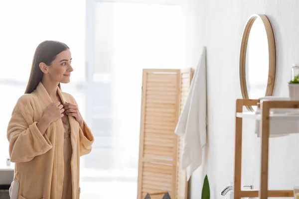 Cheerful young woman adjusting bathrobe and looking at mirror and smiling in bathroom — Stock Photo