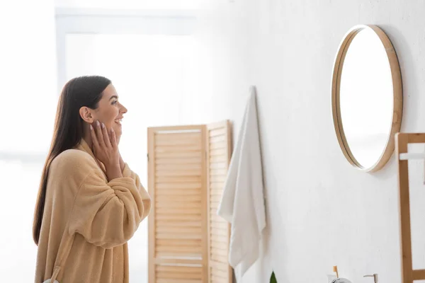 Vista laterale della donna allegra toccare la pelle pulita e guardando lo specchio in bagno — Foto stock