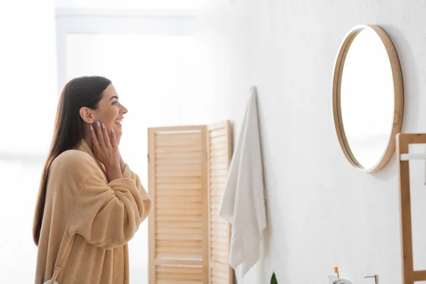 Side view of cheerful woman touching clean skin and looking at mirror in bathroom — Stock Photo