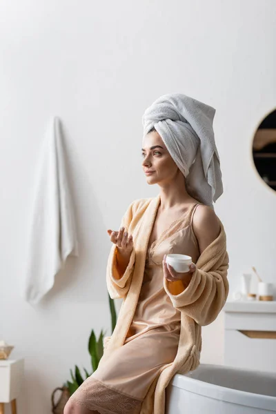 Junge Frau mit Handtuch auf dem Kopf hält Behälter mit Körperbutter im Badezimmer — Stockfoto