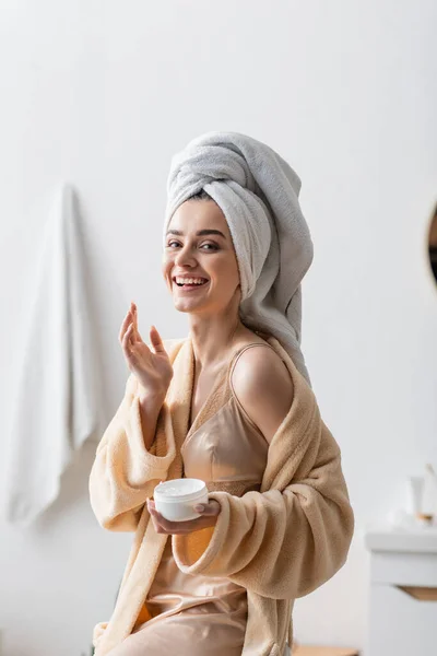 Alegre joven con toalla en la cabeza y albornoz sosteniendo contenedor con mantequilla corporal en el baño - foto de stock