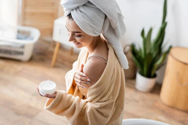 Mulher sorridente em roupão de banho com toalha na cabeça segurando recipiente com manteiga corporal — Fotografia de Stock