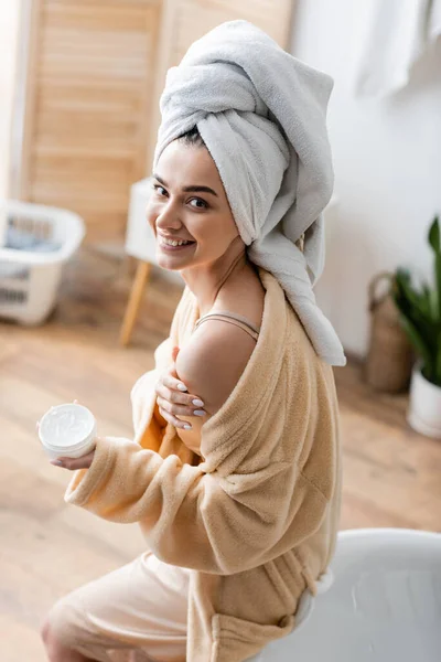 Mujer joven sonriente en albornoz con toalla en la cabeza sosteniendo contenedor con mantequilla corporal - foto de stock