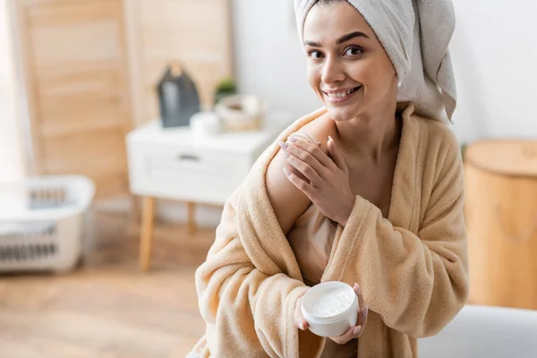 Mujer joven feliz en albornoz con toalla en el recipiente de retención de la cabeza mientras se aplica mantequilla corporal - foto de stock