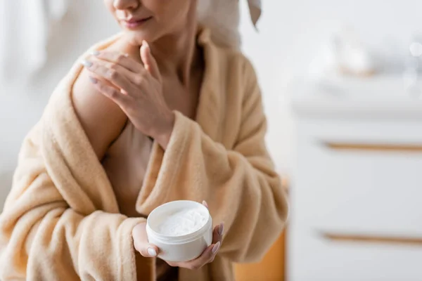 Cropped view of young woman in bathrobe applying cosmetic cream on shoulder in bathroom — Stock Photo