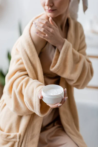 Cropped view of blurred woman in bathrobe holding cosmetic cream in bathroom — Stock Photo
