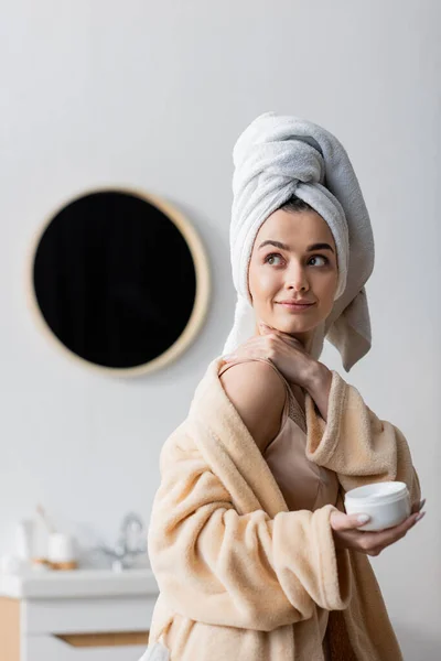 Donna sorridente in asciugamano e accappatoio applicare crema cosmetica in bagno — Foto stock
