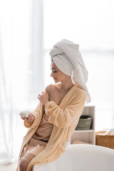 Side view of smiling woman in towel and bathrobe applying cosmetic cream near bathtub at home — Stock Photo