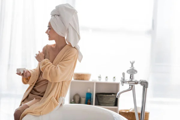 Side view of young woman in bathrobe and towel holding cosmetic cream near bathtub in bathroom — Fotografia de Stock