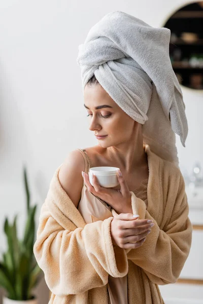 Young woman in towel on head and bathrobe holding cosmetic cream — Stock Photo