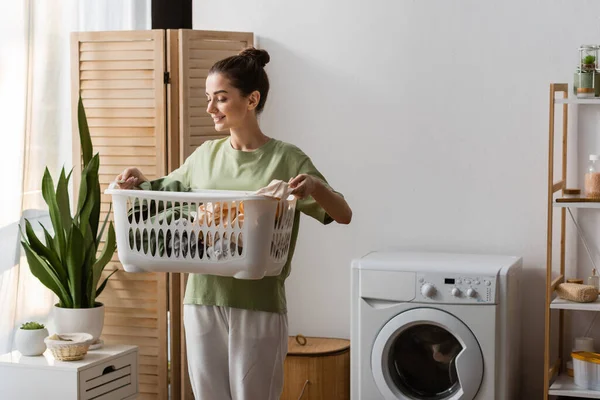 Lächelnde junge Frau betrachtet Kleidungsstücke in Wäschekorb in der Waschküche — Stockfoto