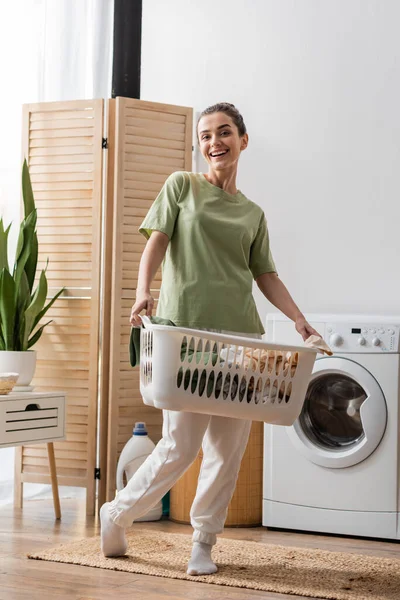 Cheerful young woman holding basket with dirty clothes near washing machine at home — Stock Photo