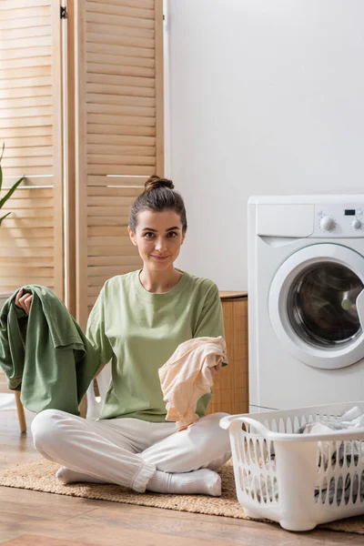 Positive Frau hält Kleidung in der Nähe von Korb und Waschmaschine zu Hause — Stockfoto