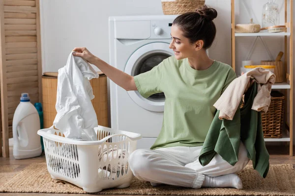 Glückliche junge Frau sortiert Kleidung neben Waschmaschine in Waschküche — Stockfoto