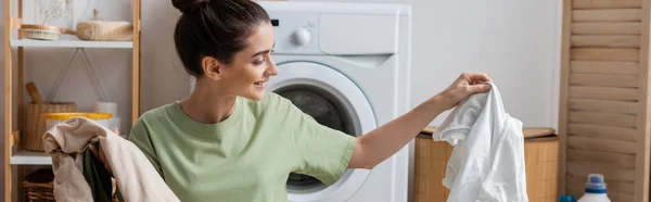 Mujer sonriente sosteniendo ropa blanca y colorida en la sala de lavandería, pancarta - foto de stock