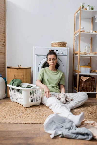 Mujer cansada sentada cerca de la ropa y la lavadora en la lavandería - foto de stock