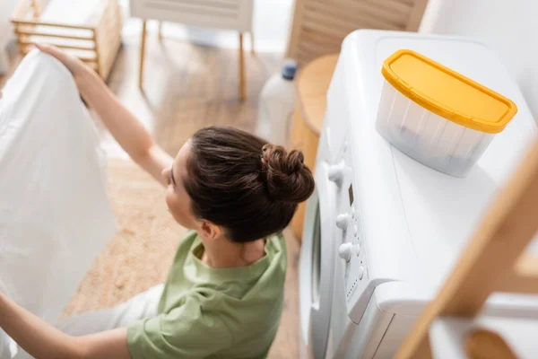 Vue aérienne de la femme brune tenant des vêtements propres près de la machine à laver dans la buanderie — Photo de stock