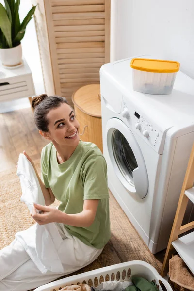Вид улыбающейся женщины, смотрящей на коробку на стиральной машине рядом с корзиной с одеждой в стиральной комнате — стоковое фото