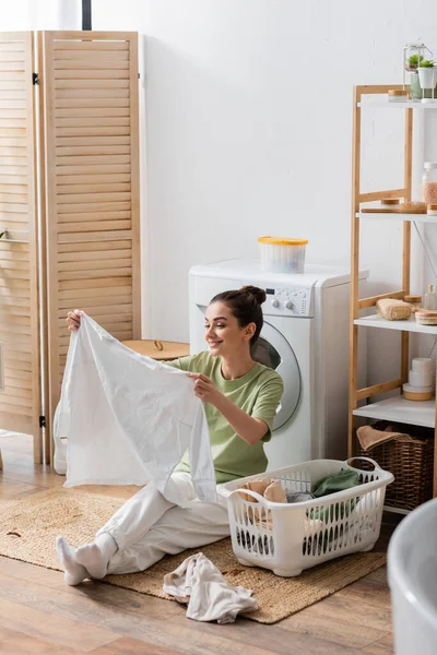 Lächelnde Frau hält Hemd neben Korb und Waschmaschine zu Hause — Stockfoto